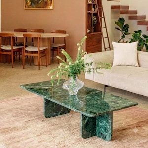 May include: A rectangular coffee table made of polished green marble with a thick slab top and two vertical slab legs. A clear glass vase containing green foliage sits in the center of the table surface, which is placed on a textured rug.