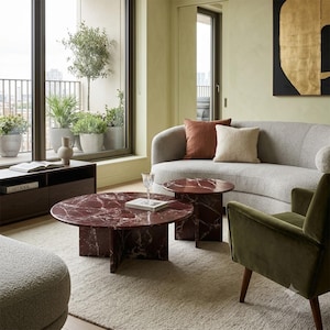May include: Two round coffee tables made of deep red marble with white veining, featuring a cross-base design. The tables are positioned in a living space with a light grey curved sofa, a green velvet armchair, and a light-coloured area rug.