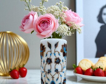 Könnte beinhalten: Eine weiße, zylindrische Vase mit einem braunen und blauen Blumenmuster enthält rosa Rosen und weiße Blüten. Die Vase steht auf einer Marmoroberfläche, mit Erdbeeren und einem goldenen Deko-Objekt im Hintergrund.
