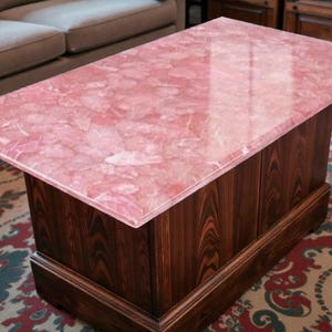 Op de afbeelding: Een rechthoekige salontafel met een roze, patroonvormig blad en een donkere houten basis. De bovenkant van de tafel heeft een gepolijst, reflecterend oppervlak. De basis is gemaakt van verticale houten panelen en een decoratieve rand.