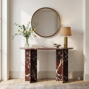 May include: A dark red marble console table with a smooth, semi-circular top and two vertical slab legs. The table is topped with a clear glass vase containing green eucalyptus and yellow flowers, two stacked books, and a small ceramic bowl.