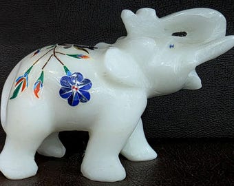 Puede incluir: Una figura de elefante de mármol blanco con incrustaciones florales. El elefante tiene una trompa levantada y está decorado con una flor azul y otros acentos florales coloridos. La figura es un objeto decorativo.
