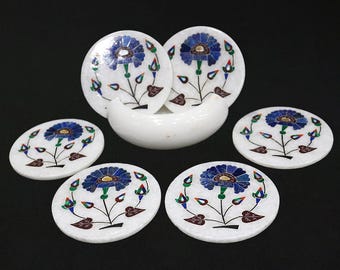 Può includere: Un set di sottobicchieri in marmo bianco con disegni floreali intarsiati. Ogni sottobicchiere presenta un fiore blu con dettagli verdi e rossi e foglie marroni. È incluso anche un supporto a forma di mezzaluna in marmo bianco.