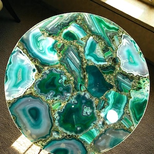 May include: A circular table featuring a mosaic top composed of green, teal, and grey agate slices. The agate is interspersed with a gold-coloured material. The table has a dark metal base. The table is set against a neutral background.