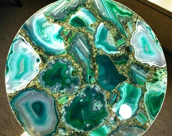 May include: A round table with a mosaic top made of green, teal, and gray agate slices. The agate pieces are interspersed with a gold-colored material. The table has a dark metal base and is set against a neutral background.