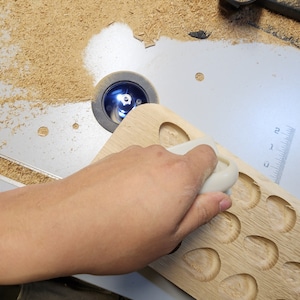May include: A woodworking project in progress. A light-colored wooden board with carved oval indentations is being shaped with a router. The router is in use, and wood shavings are visible around the work area.
