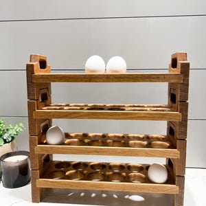 May include: A wooden egg holder with four shelves, each with 12 egg slots. The holder is made of natural wood and has a rustic look. There are three white eggs on the top shelf.