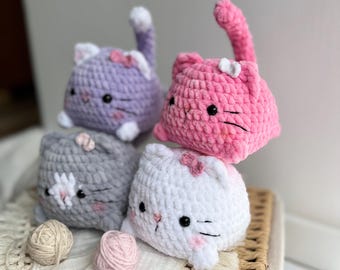 May include: Four crocheted cat plushies in various colors: purple, pink, gray, and white. Each cat has a small bow, black eyes, and pink cheeks. The cats are arranged on a woven surface with yarn balls.