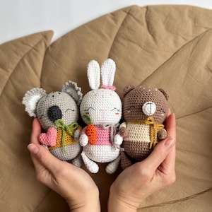 May include: Three crocheted toy animals: a koala, a bunny, and a bear. The koala is grey with a pink heart and yellow bow. The bunny is white with a pink bow and holds a carrot. The bear is brown with a yellow bow.