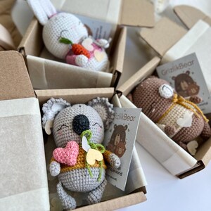 May include: Handmade crochet animal toys, including a white bunny with a carrot, a grey koala with a heart, and a brown bear, are presented in open cardboard boxes. Each toy has a unique design and a small card.