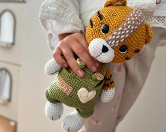 May include: A handmade crochet tiger doll with a yellow and black striped head, white muzzle, and black eyes. The tiger wears green overalls with a heart-shaped patch and the name "Dylan" on a tag.