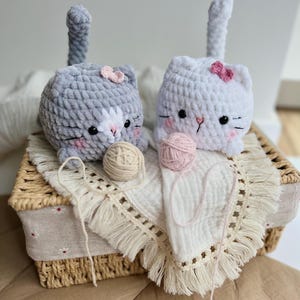 Könnte beinhalten: Zwei gehäkelte Katzenspielzeuge, eines grau und eines weiß, jeweils mit einer rosa Schleife und einem Garnknäuel. Die Katzen haben schwarze Augen und rosa Wangen. Sie liegen auf einem weißen Stoff mit Fransen in einem geflochtenen Korb.
