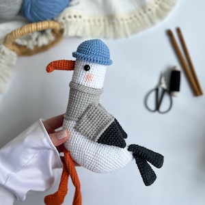 Può includere: Un giocattolo fatto a mano all'uncinetto a forma di gabbiano. Il gabbiano è bianco con ali nere e una sciarpa grigia. Ha un becco e zampe arancioni e indossa un cappello blu. Il giocattolo è alto circa 20 cm.