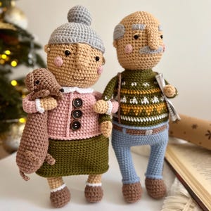 May include: Two crocheted dolls, one with a grey hat and a pink sweater, holding a brown crocheted dog, and the other with a green sweater and blue trousers.