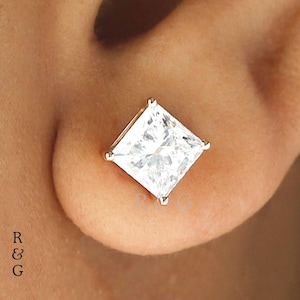 May include: A square-cut, clear gemstone stud earring. The earring is set in a silver-colored metal and is shown on an ear. The gemstone has a brilliant, faceted appearance, reflecting light. The earring is a classic jewelry piece.