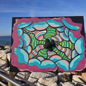 May include: A colorful abstract painting on a black canvas. The artwork features a central black shape surrounded by concentric circles of rainbow colors. Teal cloud-like shapes are outlined in pink, with a pink border.