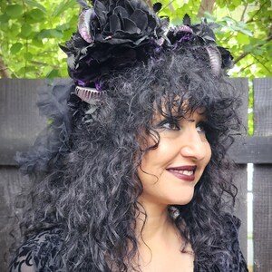 May include: A black floral headpiece with purple accents and silver embellishments. The headpiece features large, dark flowers and is adorned with feathers and decorative elements. The woman in the image is wearing a black patterned top.