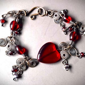 May include: A handmade bracelet featuring a large, heart-shaped red bead at the center. The bracelet is crafted with silver-colored wire, adorned with red beads and decorative swirls, and has a bronze-colored clasp.