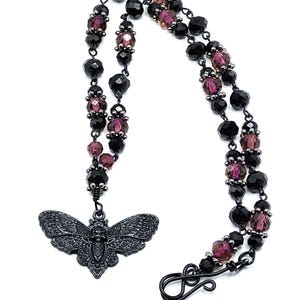 May include: A necklace featuring a dark metal moth pendant with intricate wing patterns and a central skull motif. The chain consists of alternating black and translucent purple faceted beads connected by dark metal links with a hook clasp.