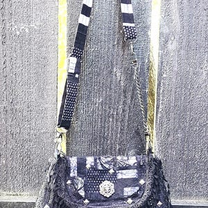 May include: A patchwork shoulder bag with a black and white color scheme. The bag features a patterned strap with stripes and polka dots, a decorative clasp, and a fringed edge. The bag is adorned with silver studs and a decorative brooch.