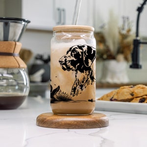 May include: A glass jar with a black and white illustration of a dog on it. The jar is filled with a brown liquid and has a straw sticking out of it. The jar is sitting on a wooden coaster.