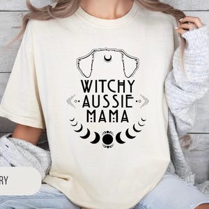 May include: Ivory-colored t-shirt with the text "Witchy Aussie Mama" in black. The design includes a dog silhouette, crescent moons, and other celestial elements. The shirt is a comfortable, casual style.