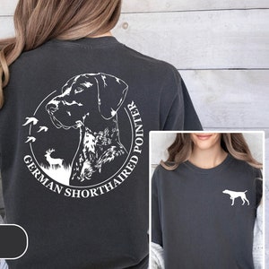 May include: Dark gray t-shirt featuring a white graphic of a German Shorthaired Pointer dog within a circular design, with the text "GERMAN SHORTHAIRED POINTER". A smaller image shows a white dog silhouette on the chest. The shirt is labeled "PEPPER".