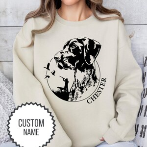 May include: A beige sweatshirt with a black and white illustration of a German Shorthaired Pointer dog with the name "Chester" printed below. The dog is looking to the left and there are three birds flying in the background. The sweatshirt has a round neckline and long sleeves.