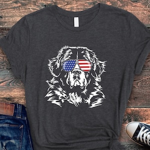 May include: Dark gray t-shirt featuring a white illustration of a dog wearing sunglasses with an American flag design. The shirt is paired with blue jeans and black and white sneakers, set against a wooden background.