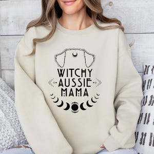 May include: Beige sweatshirt with the text "Witchy Aussie Mama" in black. The design includes a dog silhouette, arrows, and moon phases. The sweatshirt is a casual, comfortable style.