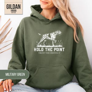 May include: A military green hooded sweatshirt featuring a white graphic of a pointing dog standing in tall grass. Below the dog, the text reads HOLD THE POINT and TRUST THE INSTINCT. The garment includes a front kangaroo pocket and a drawstring hood.