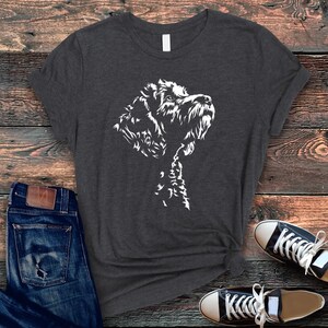 May include: Dark gray t-shirt featuring a white graphic of a dog's head and neck. The dog is depicted in a detailed, artistic style, with a focus on its fur and facial features. The shirt is paired with blue jeans and black and white sneakers.