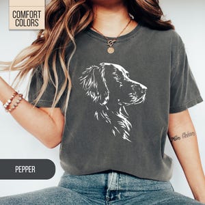 May include: Dark grey t-shirt with a white graphic of a dog's head in profile. The shirt has the text "COMFORT COLORS" at the top left and "PEPPER" at the bottom. The shirt is a crew neck and has short sleeves.