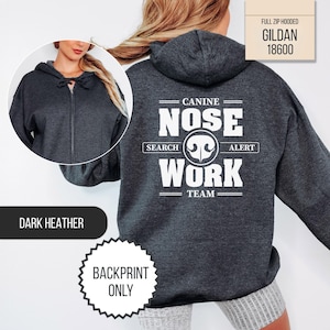 May include: A dark heather gray full-zip hooded sweatshirt featuring a white graphic on the back that reads CANINE NOSE WORK SEARCH ALERT TEAM with a dog nose icon. The front view shows a matching dark heather gray zip-up hoodie by Gildan 18600.