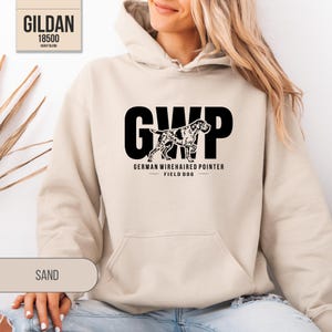 May include: A sand-colored hooded sweatshirt featuring a black graphic on the chest. The graphic displays the letters GWP, a silhouette of a German Wirehaired Pointer, and the text GERMAN WIREHAIRED POINTER FIELD DOG. A Gildan 18500 label is visible.