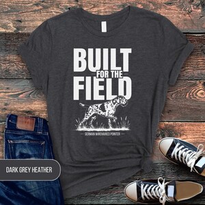 May include: A dark grey heather t-shirt featuring the white text BUILT FOR THE FIELD above a graphic illustration of a German Wirehaired Pointer dog standing in grass. The text GERMAN WIREHAIRED POINTER is printed in white below the dog illustration.