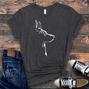 May include: Dark gray t-shirt featuring a white line drawing of a dog's profile. The dog's head and neck are depicted in a minimalist style. The shirt is paired with blue jeans and black and white sneakers, creating a casual, everyday look.