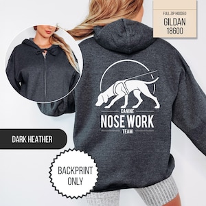 May include: A dark heather gray full-zip hooded sweatshirt featuring a white graphic on the back of a dog in a harness with the text CANINE NOSE WORK TEAM. The front view shows a matching dark heather gray zip-up hoodie with a drawstring hood.