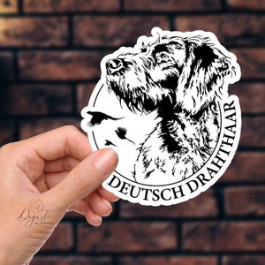 May include: A black and white sticker featuring a Deutsch Drahthaar dog with the text "DEUTSCH DRAHTHAAR" in a circular design. The sticker also includes three flying birds. The sticker is held in a hand.
