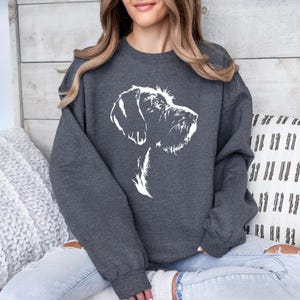 May include: Dark gray sweatshirt featuring a white line drawing of a dog's head in profile. The dog has a long snout and shaggy fur. The sweatshirt is a solid color and has long sleeves.