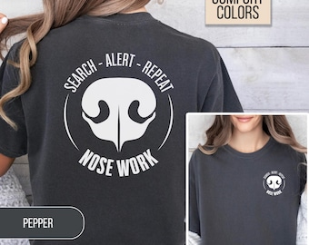 May include: A charcoal gray t-shirt featuring a white graphic on the back and a smaller version on the front. The design displays a dog nose silhouette surrounded by the text SEARCH - ALERT - REPEAT and NOSE WORK in white lettering.