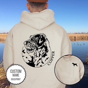 May include: A cream-colored hoodie with a black and white illustration of a dog and the word "COPPER" on the back. The hoodie has a pocket on the front and the text "CUSTOM NAME BACKPRINT" on a white circle.