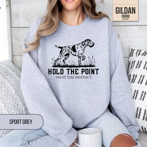 May include: A light gray crewneck sweatshirt featuring a black graphic of a hunting dog standing in grass. The text reads HOLD THE POINT and TRUST THE INSTINCT. The garment is labeled as a Gildan 18000 Heavy Blend in the color Sport Grey.