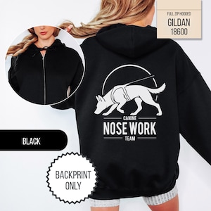 May include: A black full-zip hooded sweatshirt featuring a white graphic on the back of a dog in a harness with the text CANINE NOSE WORK TEAM. The front view shows a solid black hooded sweatshirt with a silver zipper and a drawstring hood.