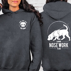May include: A dark gray hooded sweatshirt with a white graphic on the front that says "Search Alert Repeat Nose Work" and a white graphic on the back that says "Canine Nose Work Team" with a stylized illustration of a dog sniffing the ground.