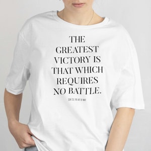 Puede incluir: Camiseta blanca con la cita "LA MAYOR VICTORIA ES LA QUE NO REQUIERE BATALLA." en texto negro. La cita es de Sun Tzu, El Arte de la Guerra. La camiseta es de estilo casual.