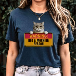 May include: Navy blue t-shirt featuring a gray cat peeking over a sign. The sign reads "WARNING: NOT A MORNING PERSON" in white text on a red and yellow background. The cat has yellow eyes and is looking forward.