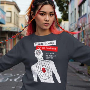 May include: Dark gray sweatshirt with the text "I used to miss my ex husband, but now my aim is better" above a graphic of a target with a person's head as the bullseye. The text is in white and red.