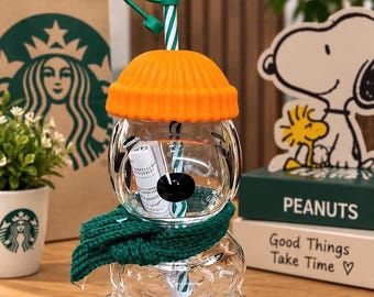 May include: A clear glass Starbucks x Peanuts tumbler shaped like a bear, wearing a bright orange ribbed beanie and a dark green knit scarf. The tumbler includes a green and white striped straw topped with a yellow Woodstock figure.