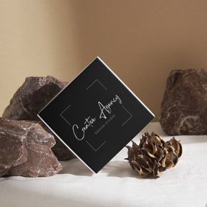 May include: A black square business card with silver lettering that reads "Creative Agency Design Studio" on a white background. The card is resting on a white surface with brown rocks and a dried flower.
